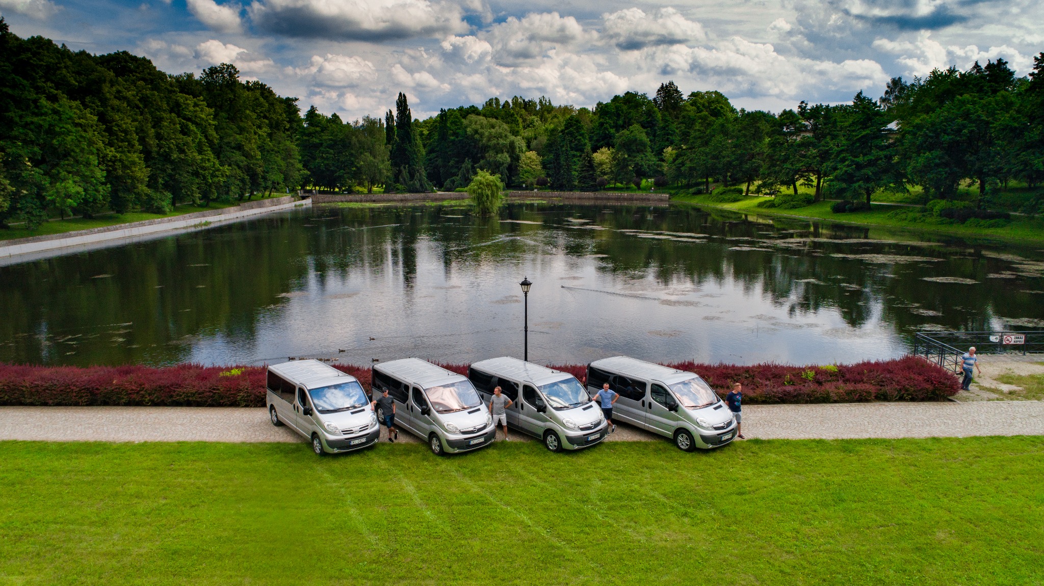 Cztery srebrne busy zaparkowane na trawie nad brzegiem jeziora, z widocznymi osobami stojącymi przy pojazdach, odbicie chmur w szybach, park w tle.