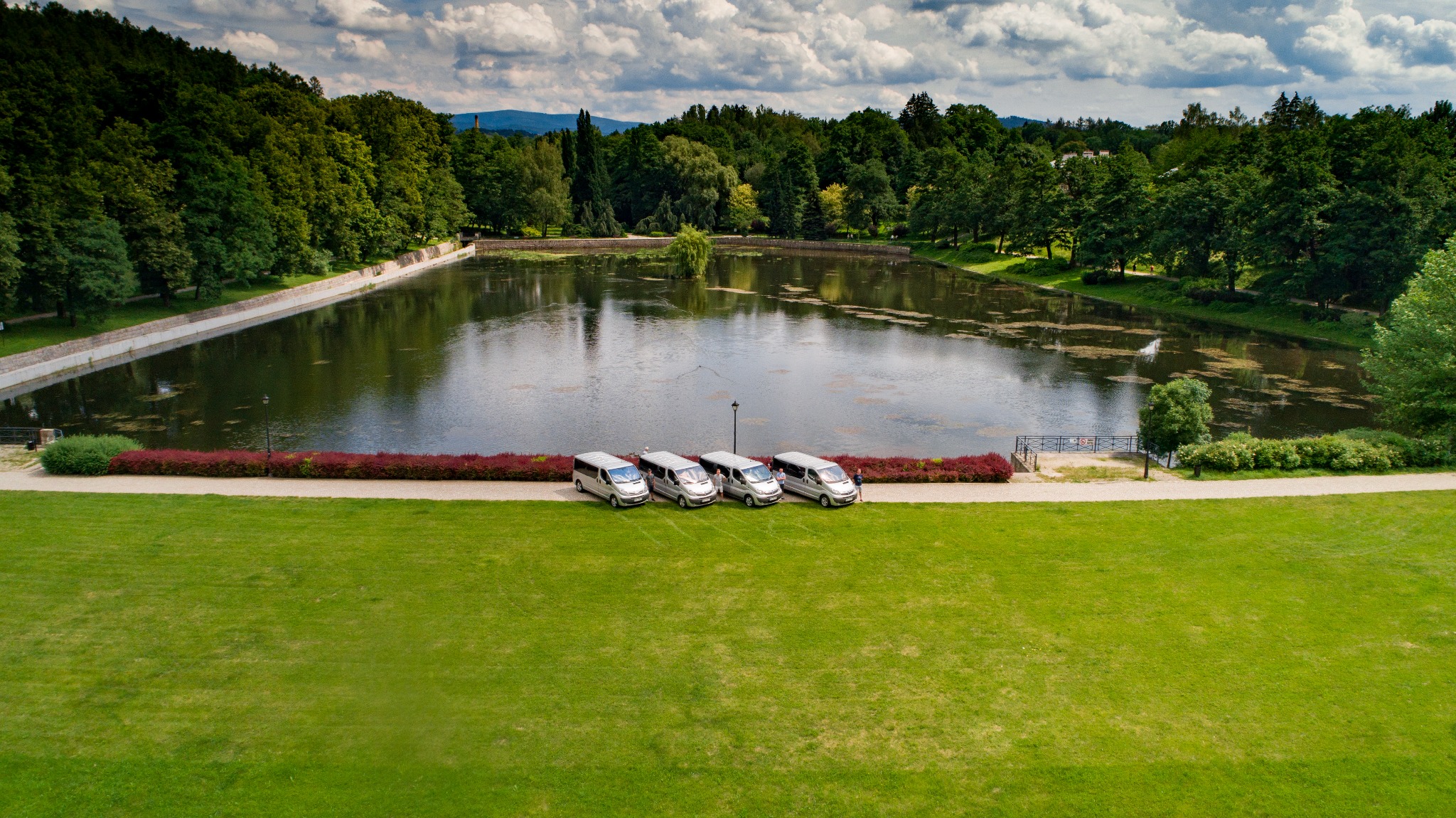 Cztery srebrne busy ustawione rzędem na trawniku przed jeziorem otoczonym zielenią, widok z góry.