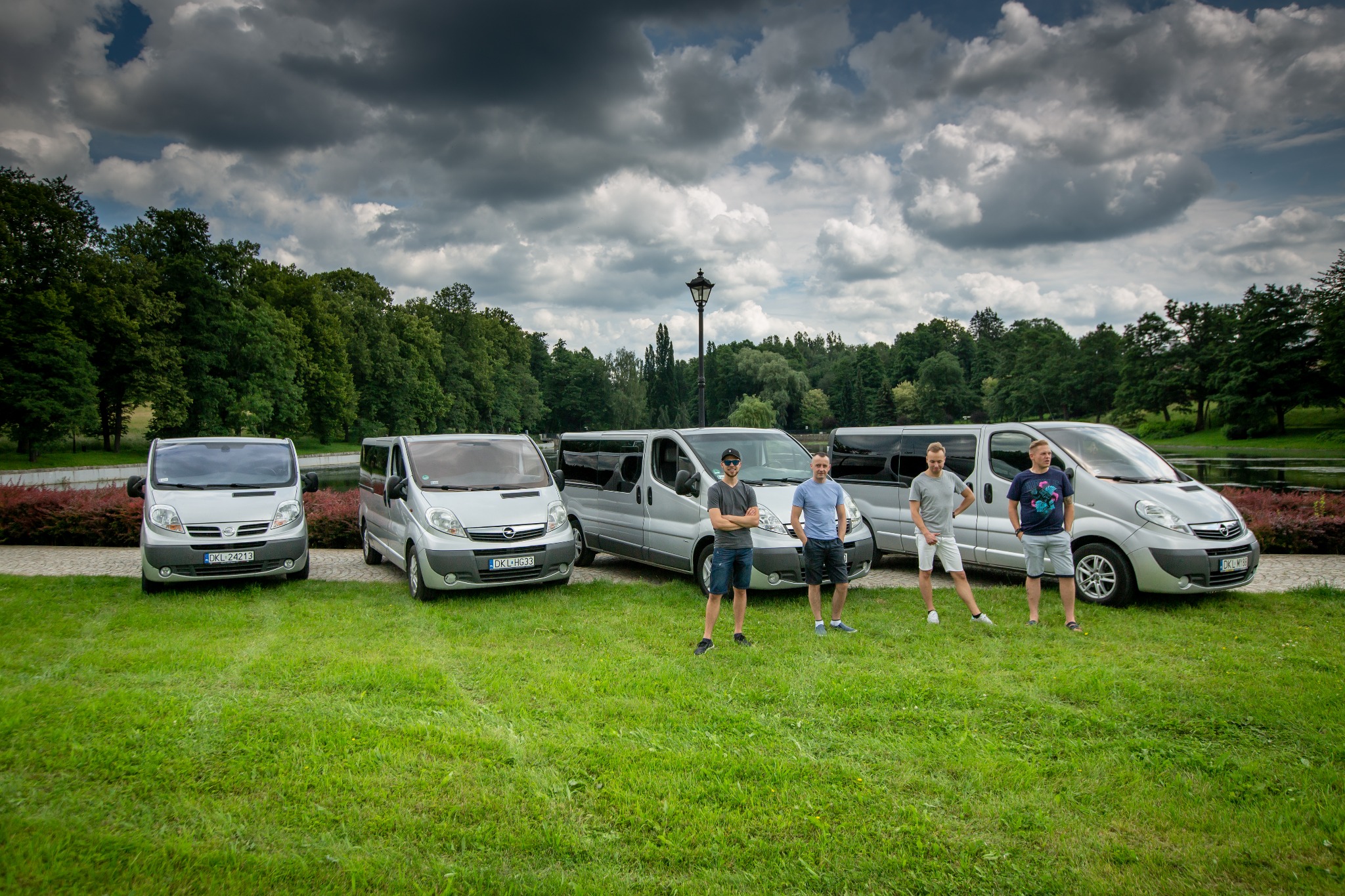 Flota srebrnych vanów Opel Vivaro na trawniku przed parkiem, z czterema mężczyznami stojącymi przed środkowymi pojazdami.