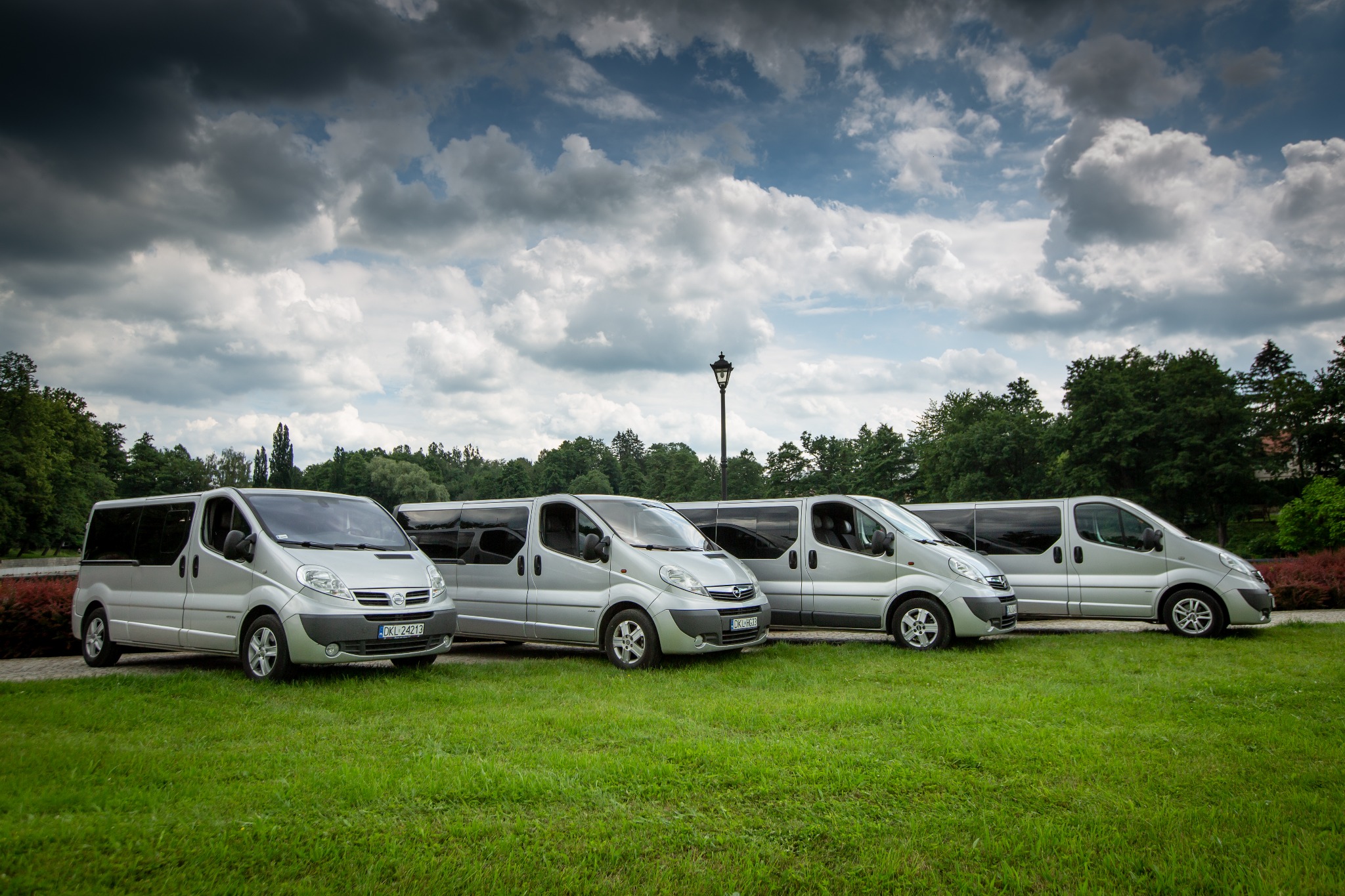 Cztery srebrne busy Opel Vivaro zaparkowane w rzędzie na trawniku, z widocznymi numerami rejestracyjnymi i parkową latarnią w tle.