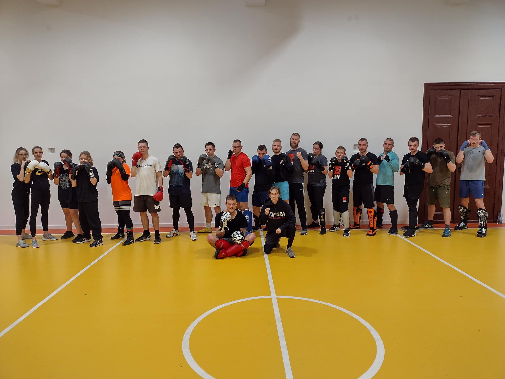 Grupa osób w rękawicach bokserskich i ochraniaczach stoi na sali z żółtą podłogą, dwóch siedzi na podłodze z trofeum i logiem Kimura na bluzie.