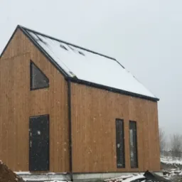 Nowoczesny, drewniany dom modułowy z dachem pokrytym śniegiem, widziany w zimowej scenerii podczas opadów śniegu.