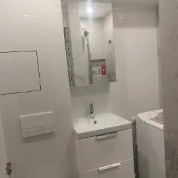 Nowoczesna łazienka w bieli: umywalka z szafką, toaleta podwieszana z przyciskiem, pralka i lustro z odbiciem kabiny prysznicowej.