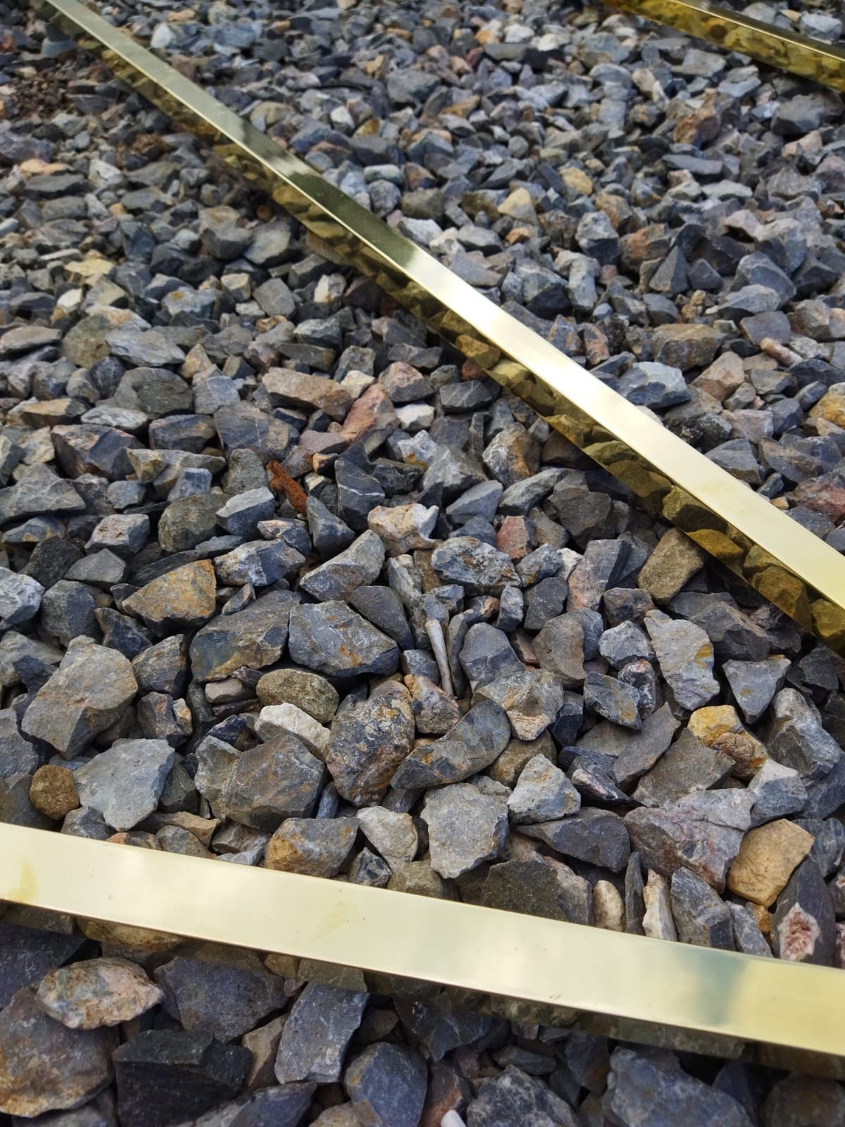 Dwa prostokątne profile z mosiądzu leżące na szarym żwirze o różnej wielkości kamieni.