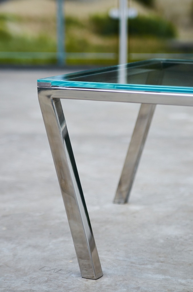Stolik kawowy z blatem ze szkła hartowanego i nogami ze stali nierdzewnej o polerowanym wykończeniu, widok z bliska na nogę z delikatnym odbiciem światła.