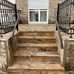 Schody zewnętrzne wyłożone płytkami imitującymi drewno, z kutymi balustradami, prowadzące do wejścia z oknem z roletą zewnętrzną, na tle kamiennej elewacji.