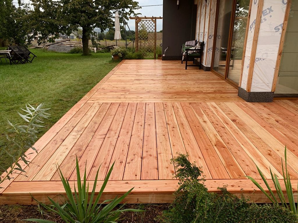 Nowoczesny taras z desek drewnianych, z widoczną strukturą drewna, przylegający do domu z dużymi oknami i zielonym trawnikiem w tle.