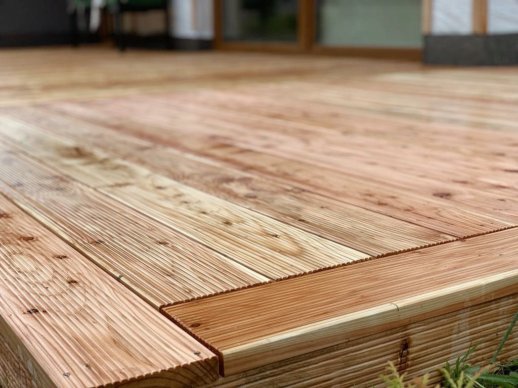 Drewniany taras z desek o jasnym odcieniu, widoczne detale łączeń i faktura drewna, trawa w dolnym rogu kadru.