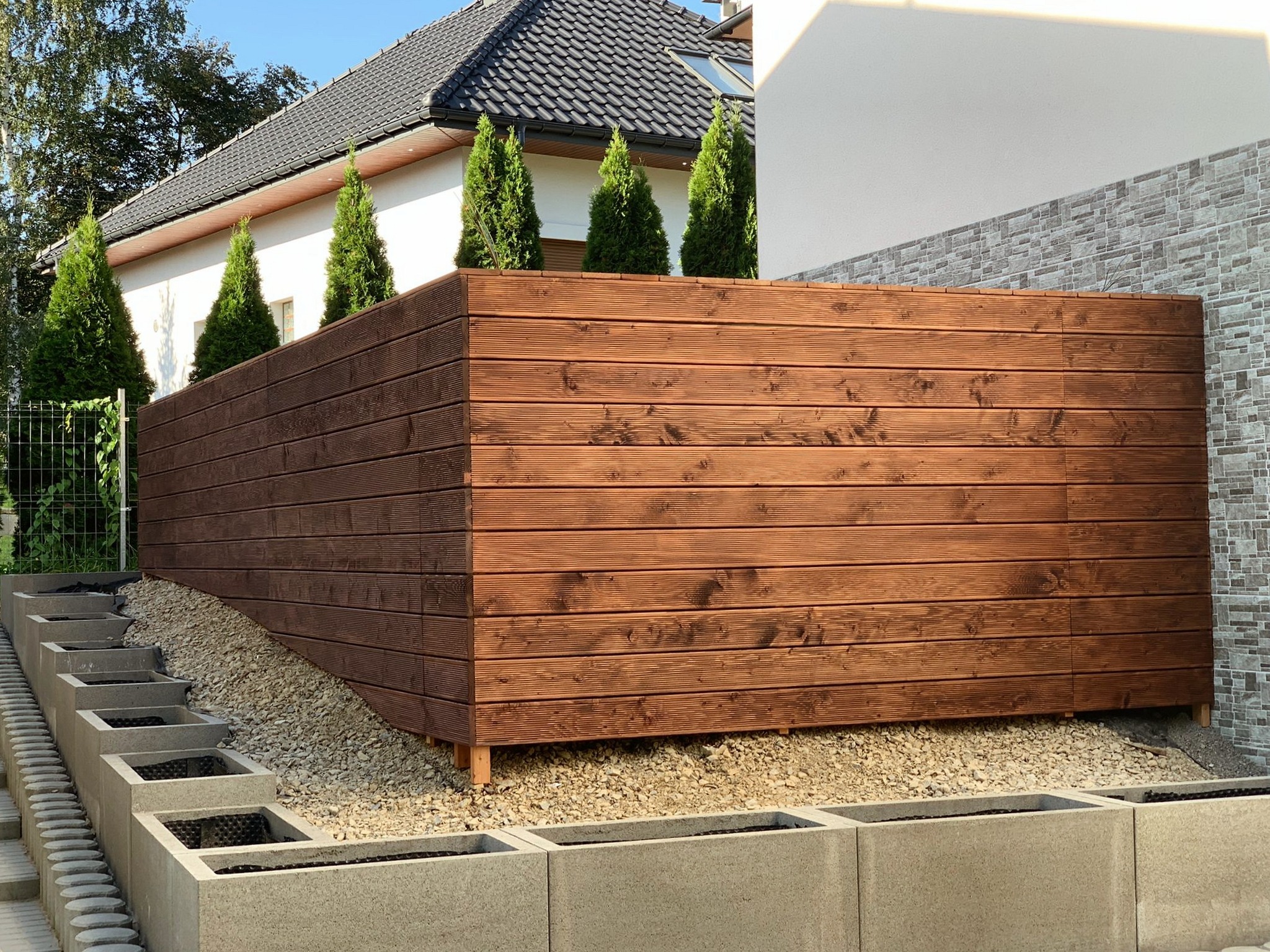 Nowoczesne ogrodzenie z ciemnego drewna, poziome deski, podwyższone na małych nóżkach, z nasypem z kamyków, obok schody z betonowych donic, w tle dom z ciemną dachówką i iglaki.
