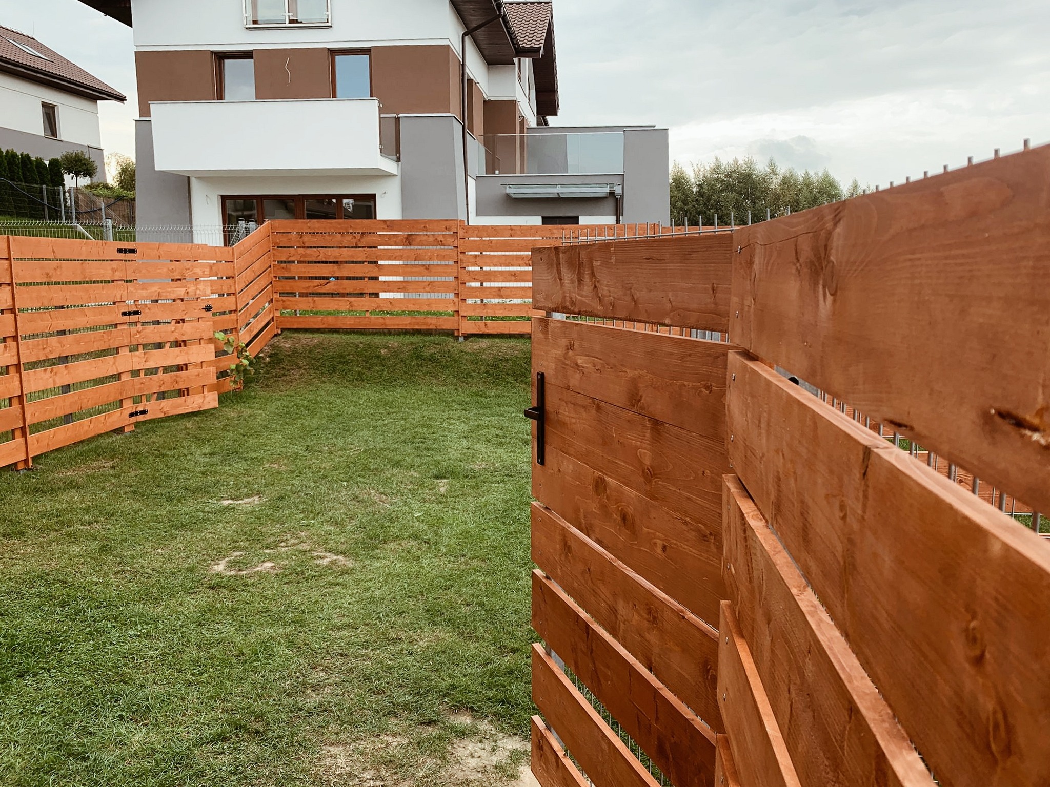 Nowoczesne ogrodzenie z drewna w kolorze miodowym, z widoczną furtką i metalowymi słupkami, na tle domu z balkonem i zielonego trawnika.