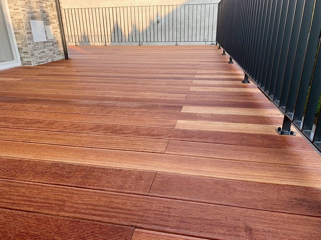 Nowoczesny taras z desek kompozytowych w odcieniach brązu, z czarną metalową balustradą i fragmentem elewacji budynku w tle.