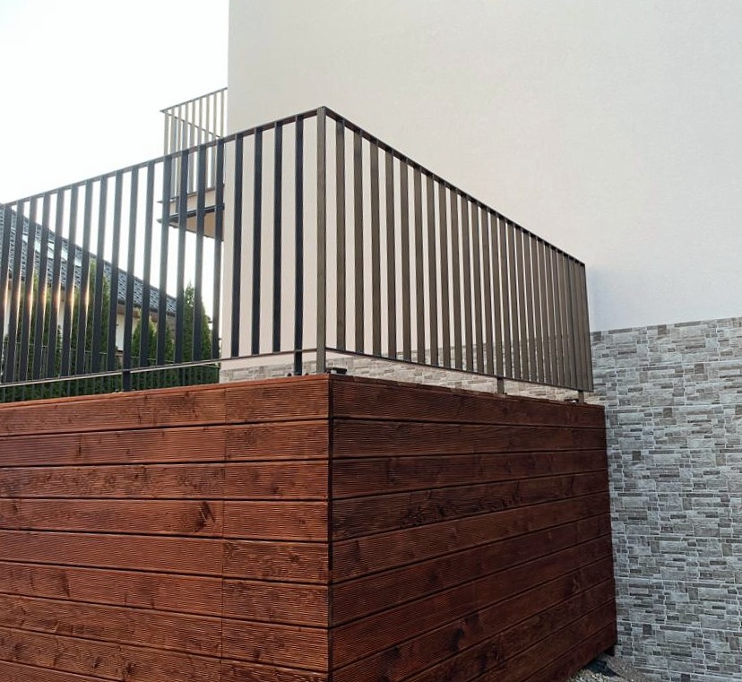 Drewniana konstrukcja tarasu z metalową balustradą, przylegająca do ściany budynku wykończonej płytkami imitującymi kamień.