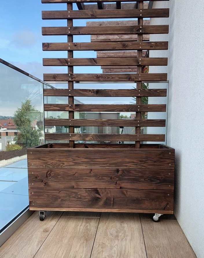Drewniana konstrukcja z poziomych listew, pełniąca funkcję osłony lub dekoracji, zamontowana w dużej donicy na kółkach, stojąca na balkonie z przeszkloną balustradą, widok na zabudowania w tle.