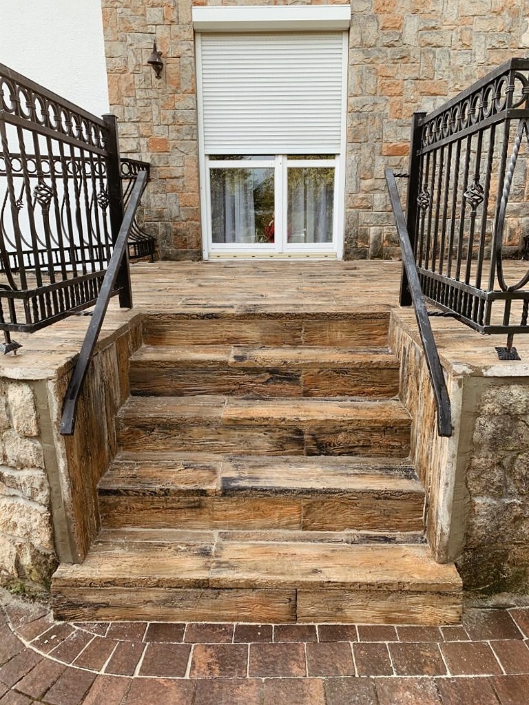 Schody zewnętrzne wyłożone płytkami imitującymi drewno, z kutymi balustradami, prowadzące do wejścia z oknem z roletą zewnętrzną, na tle kamiennej elewacji.