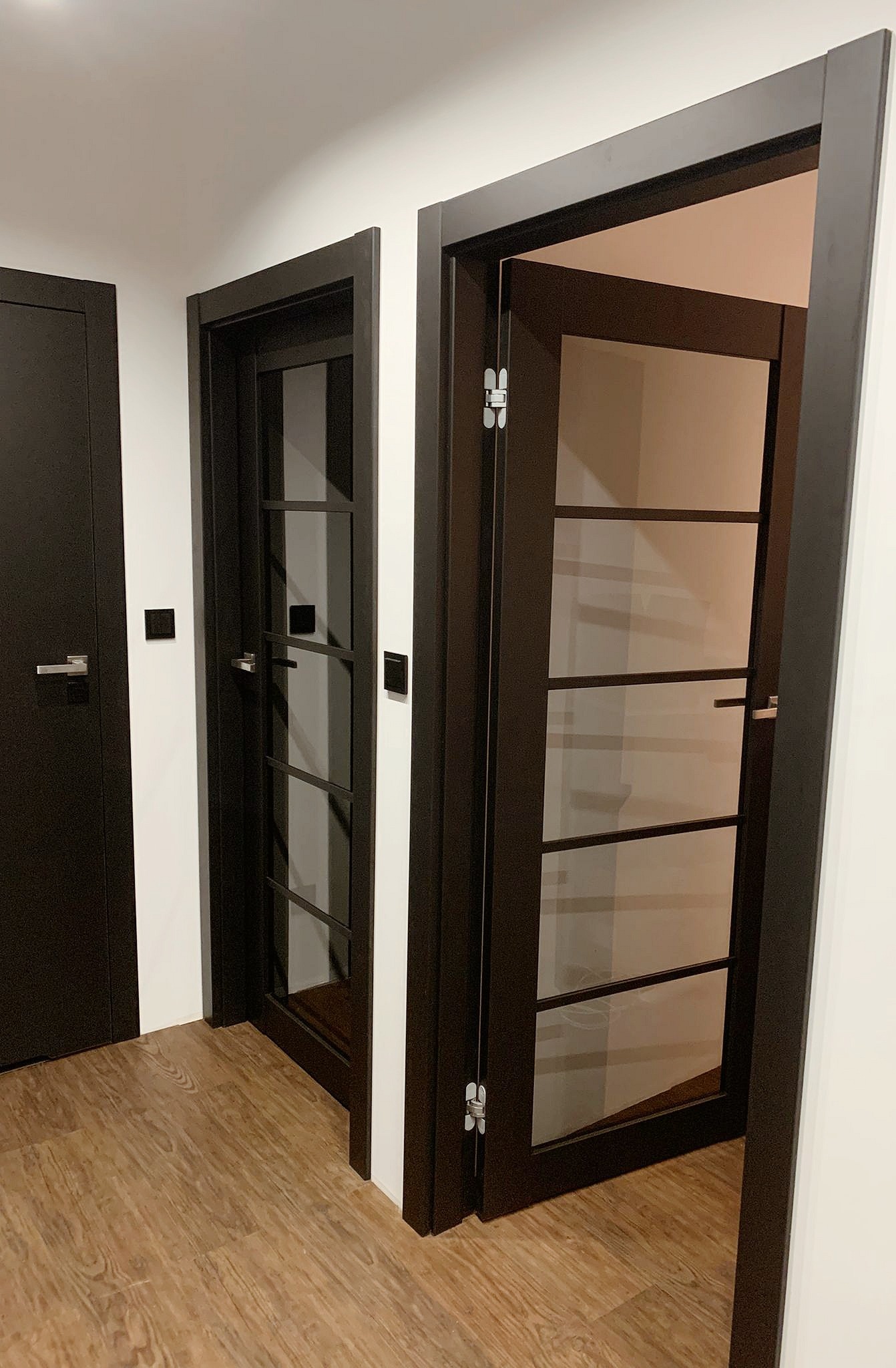 Nowoczesne, ciemne drzwi wewnętrzne z przeszkleniami, częściowo otwarte, na tle podłogi z paneli imitujących drewno.