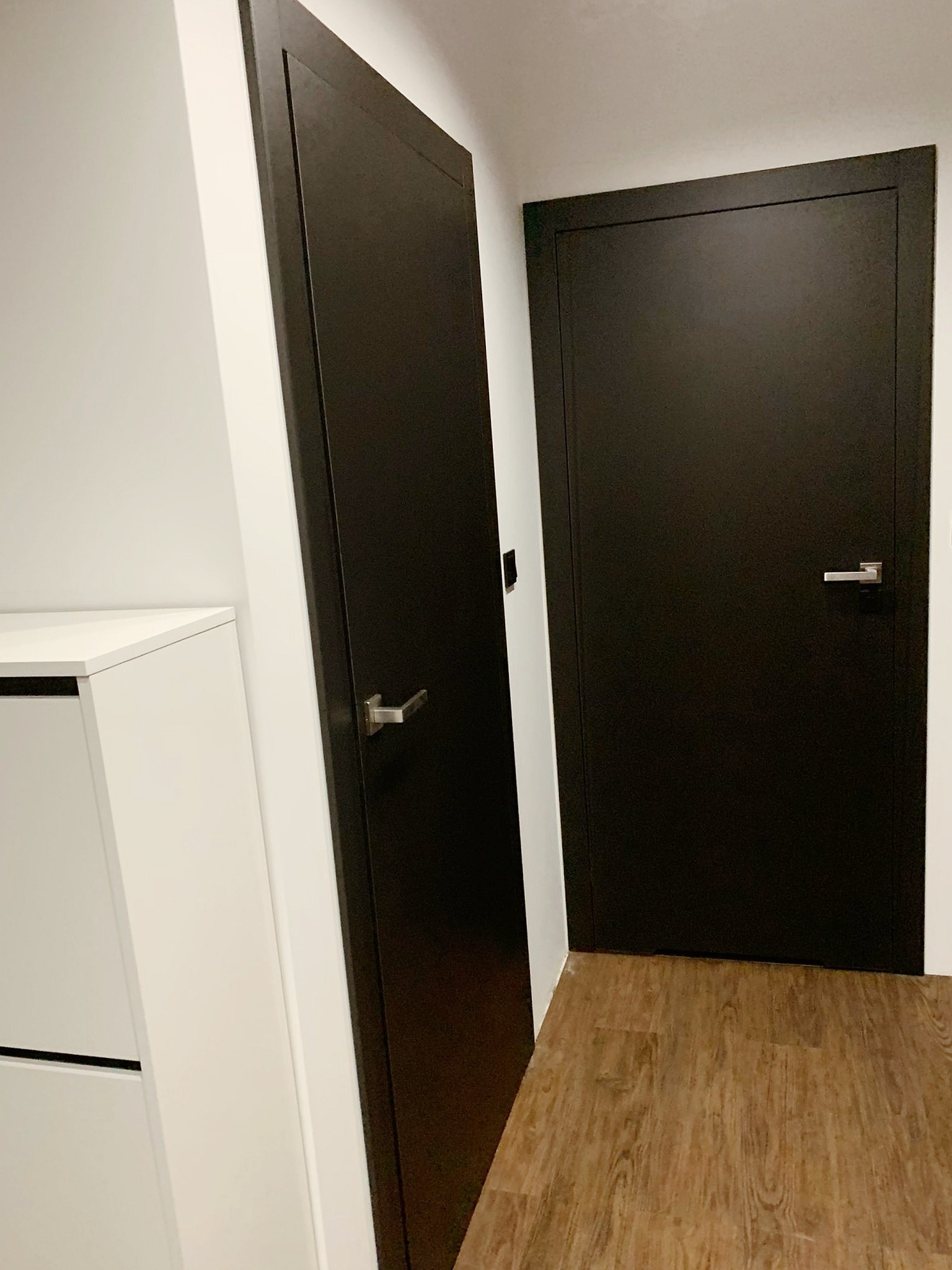 Dwie nowoczesne, ciemne drzwi wewnętrzne z metalowymi klamkami, ustawione pod kątem na tle białych ścian i podłogi w kolorze drewna. Widoczny fragment białej szafki z szufladami.