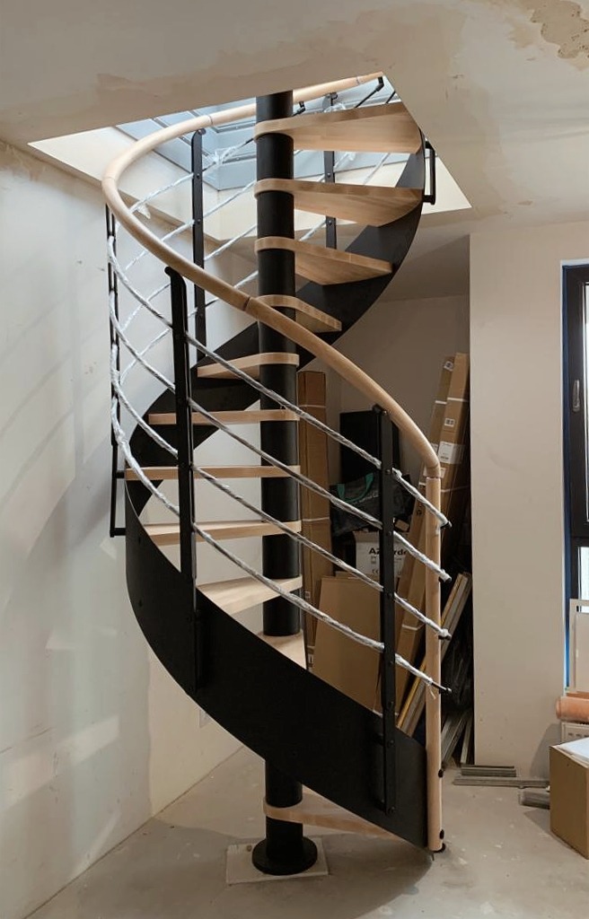Nowoczesne schody spiralne z drewnianymi stopniami i metalową konstrukcją, widoczne podczas prac wykończeniowych w pomieszczeniu z oknem.
