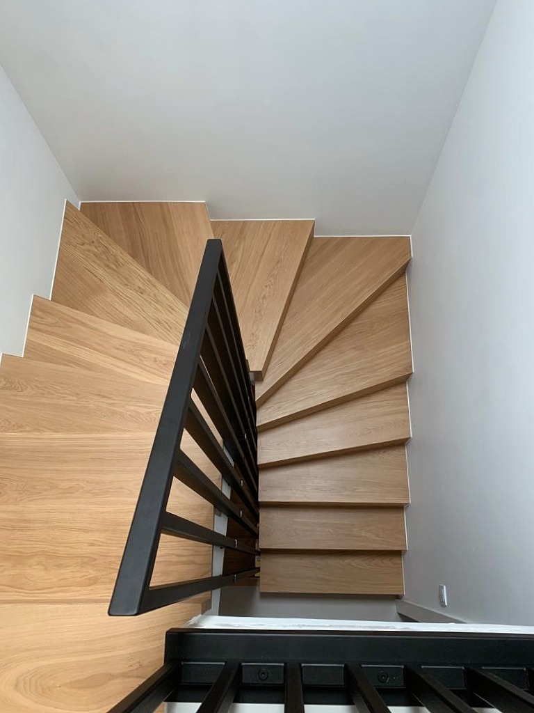 Drewniane schody spiralne z czarną, metalową balustradą, widziane z dołu, z jasnymi ścianami i sufitem.