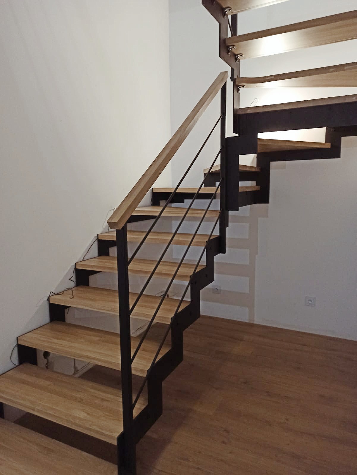 Nowoczesne schody z drewnianymi stopniami i czarną metalową konstrukcją oraz drewnianą poręczą, widoczne na tle białej ściany i drewnianej podłogi.