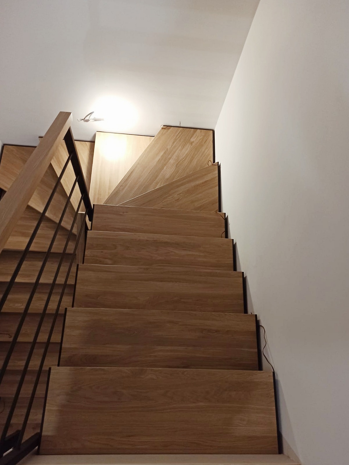 Schody drewniane o jasnym odcieniu, z czarną metalową balustradą, prowadzące do góry, oświetlone punktowym światłem na suficie.