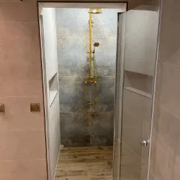 Nowoczesna kabina prysznicowa z złotą armaturą, płytki imitujące drewno na podłodze, szare płytki z wzorem na ścianie prysznica, wnęka w ścianie.