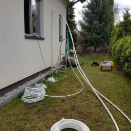 Instalacja białych rur na zewnątrz budynku, widoczny pracownik na drabinie, narzędzia i zwinięte rury na trawie.