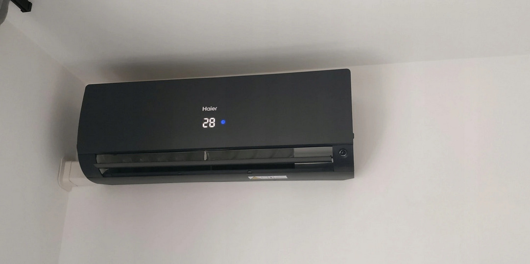 Czarny klimatyzator Haier zamontowany na białej ścianie, wyświetlający temperaturę 28 stopni Celsjusza.