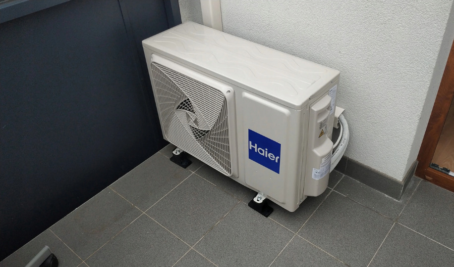 Zewnętrzna jednostka klimatyzacji Haier zamontowana na czarnych gumowych podstawkach na szarych płytkach, przy ciemnej ścianie i jasnej ścianie z oknem.
