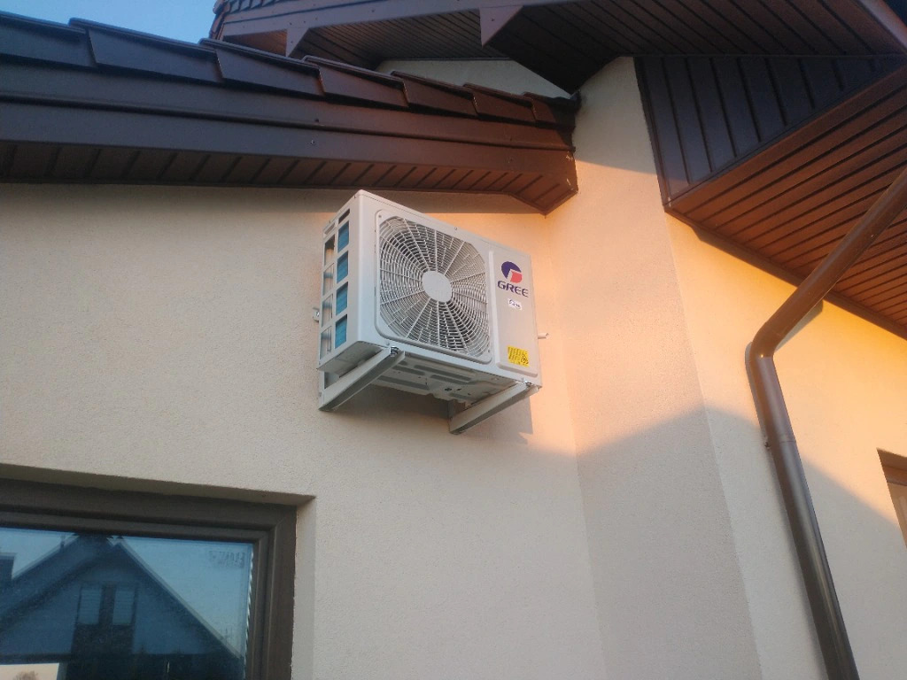 Zewnętrzna jednostka klimatyzacji marki Gree zamontowana na elewacji budynku jednorodzinnego, widoczna rynna i okno z odbiciem dachu sąsiedniego domu.