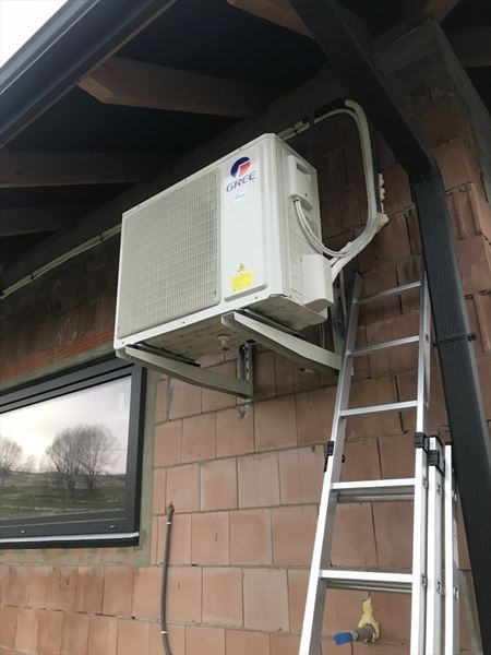 Zewnętrzna jednostka klimatyzatora marki Gree zamontowana na ścianie budynku z cegły, widoczna drabina przystawna i okno z odbiciem drzewa.