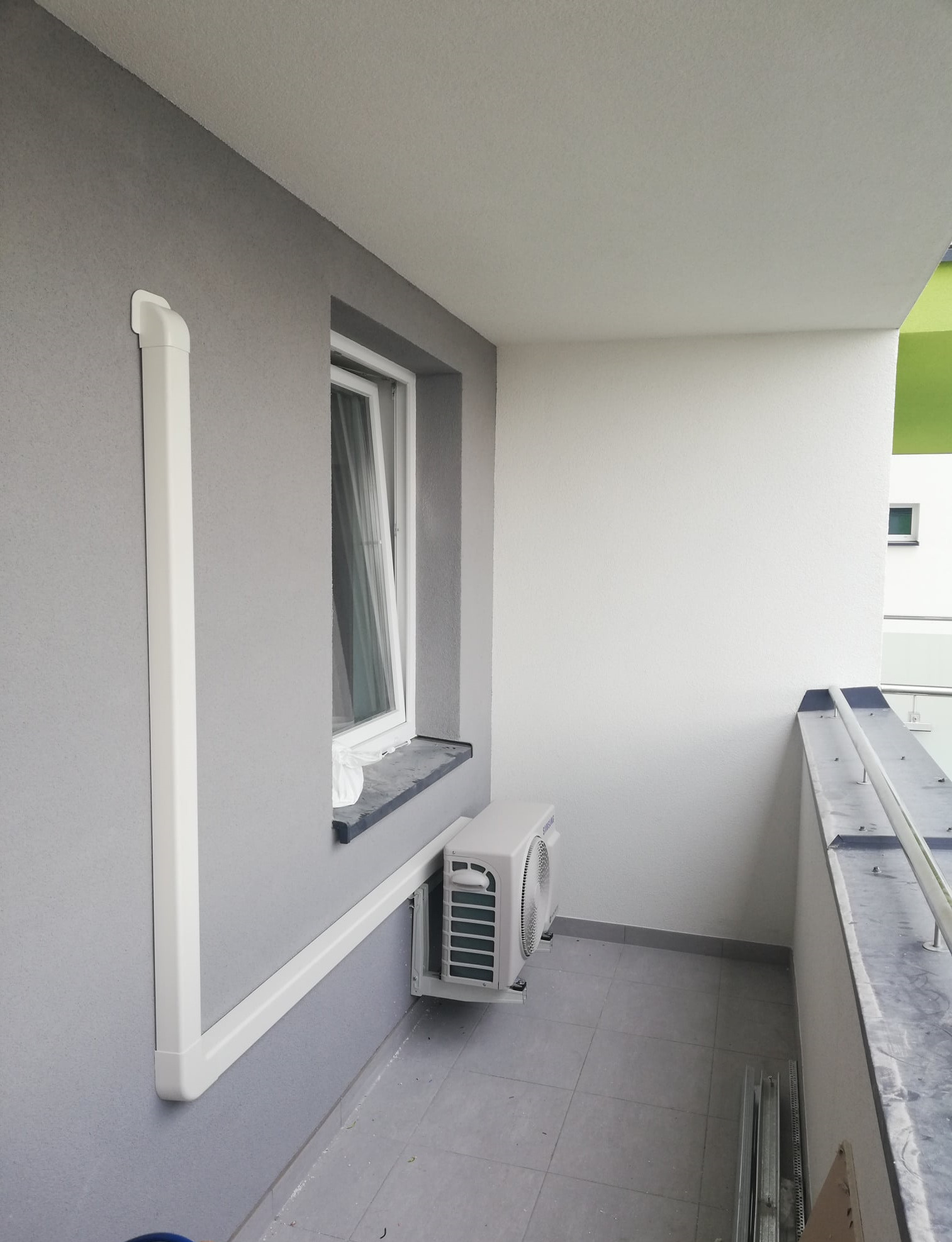 Balkon z zamontowaną jednostką zewnętrzną klimatyzatora Samsung, widoczne białe rury maskujące instalację, szare płytki na podłodze i fragment metalowej balustrady.