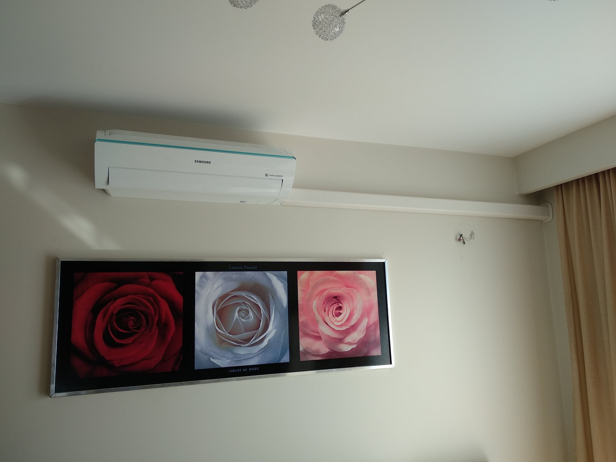 Biały klimatyzator Samsung zamontowany na ścianie nad obrazem z motywem róż, widoczne dwie lampy na suficie.