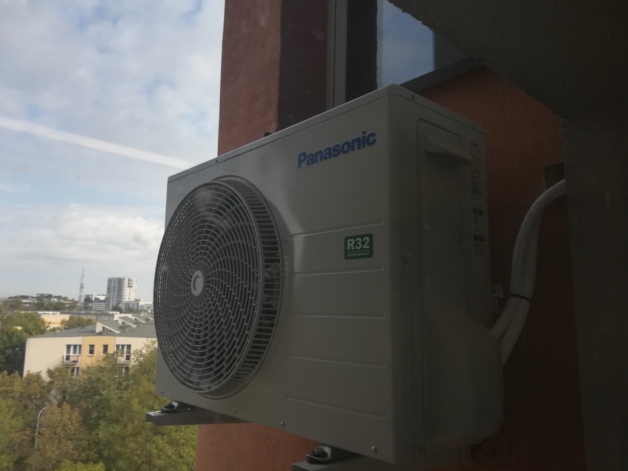 Zewnętrzna jednostka klimatyzacji Panasonic R32 zamontowana na ścianie budynku z widokiem na miasto w tle, widoczne przewody instalacyjne.