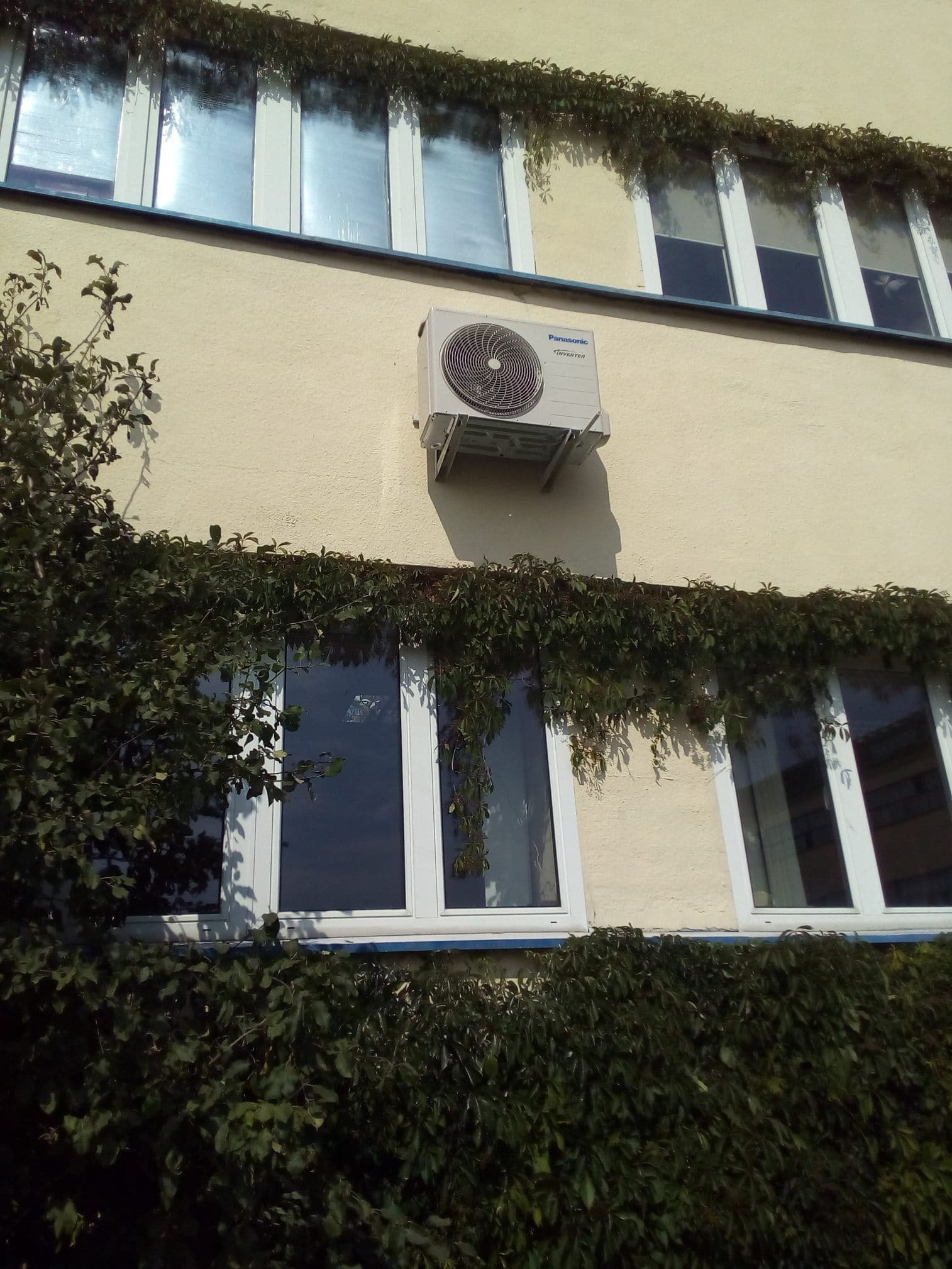 Zewnętrzna jednostka klimatyzacji Panasonic Inverter zamontowana na elewacji budynku z oknami, częściowo porośniętej bluszczem.
