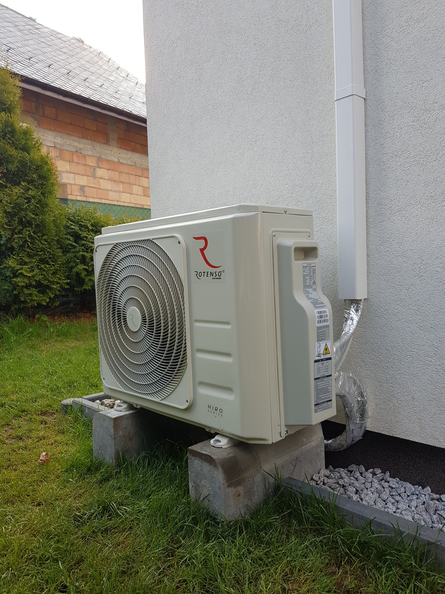 Zewnętrzna jednostka klimatyzacji Rotenso HiRO Series zamontowana na betonowych bloczkach obok ściany budynku, z widoczną izolacją rury odprowadzającej skropliny i fragmentem dachu w tle.