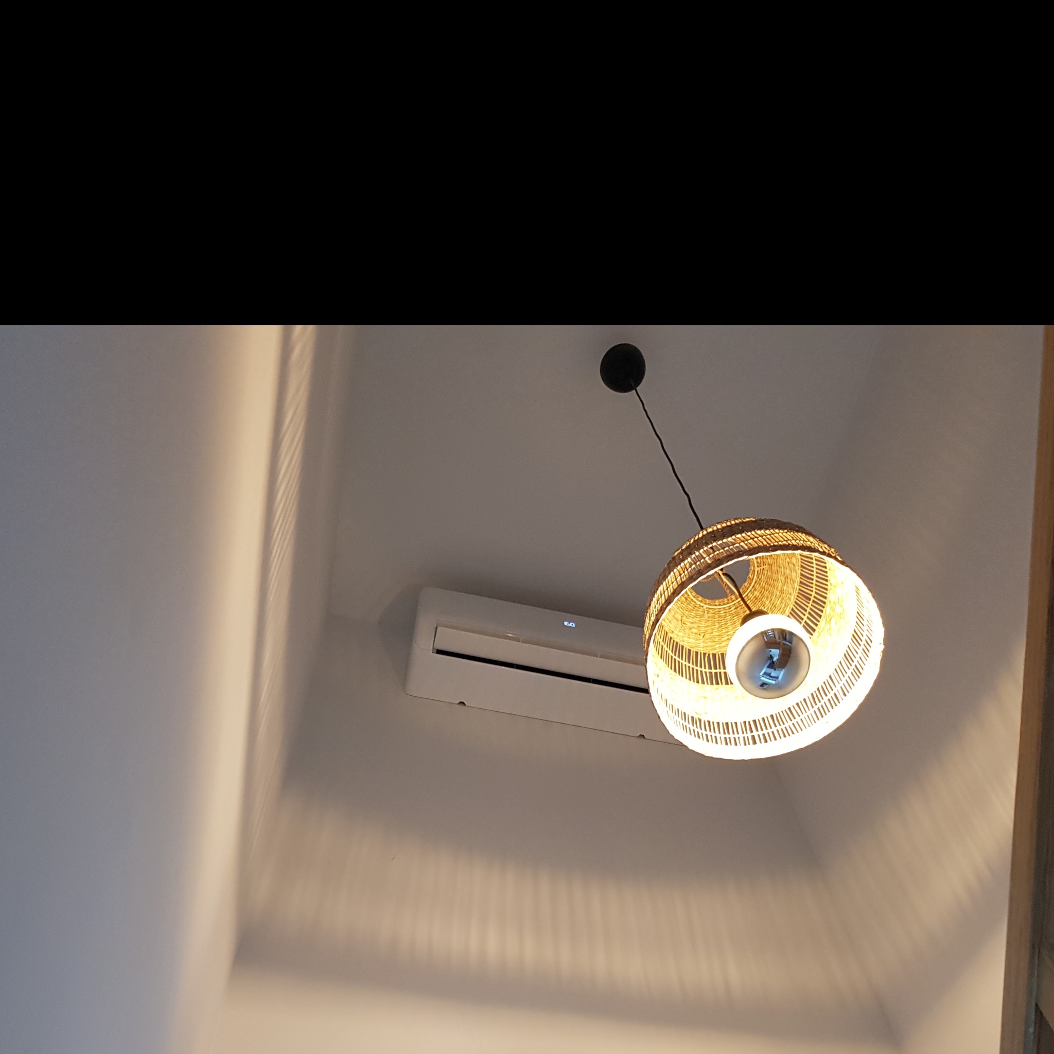 Biały klimatyzator ścienny włączony, widoczny spod ozdobnej, plecionej lampy sufitowej, rzucającej cienie na ścianę.