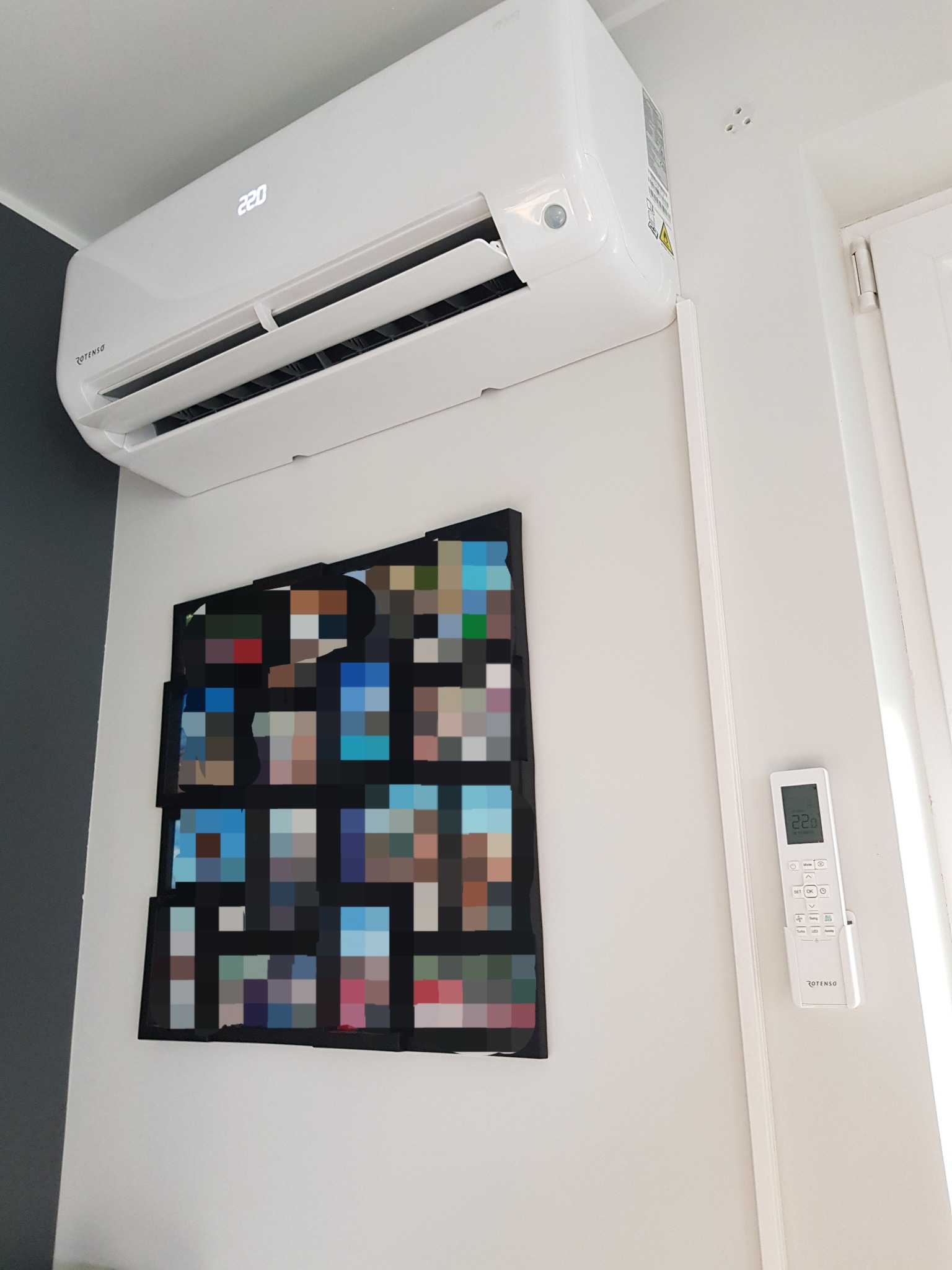 Biały klimatyzator marki Rotenso zamontowany na ścianie nad abstrakcyjnym obrazem, obok biały pilot do sterowania klimatyzacją.