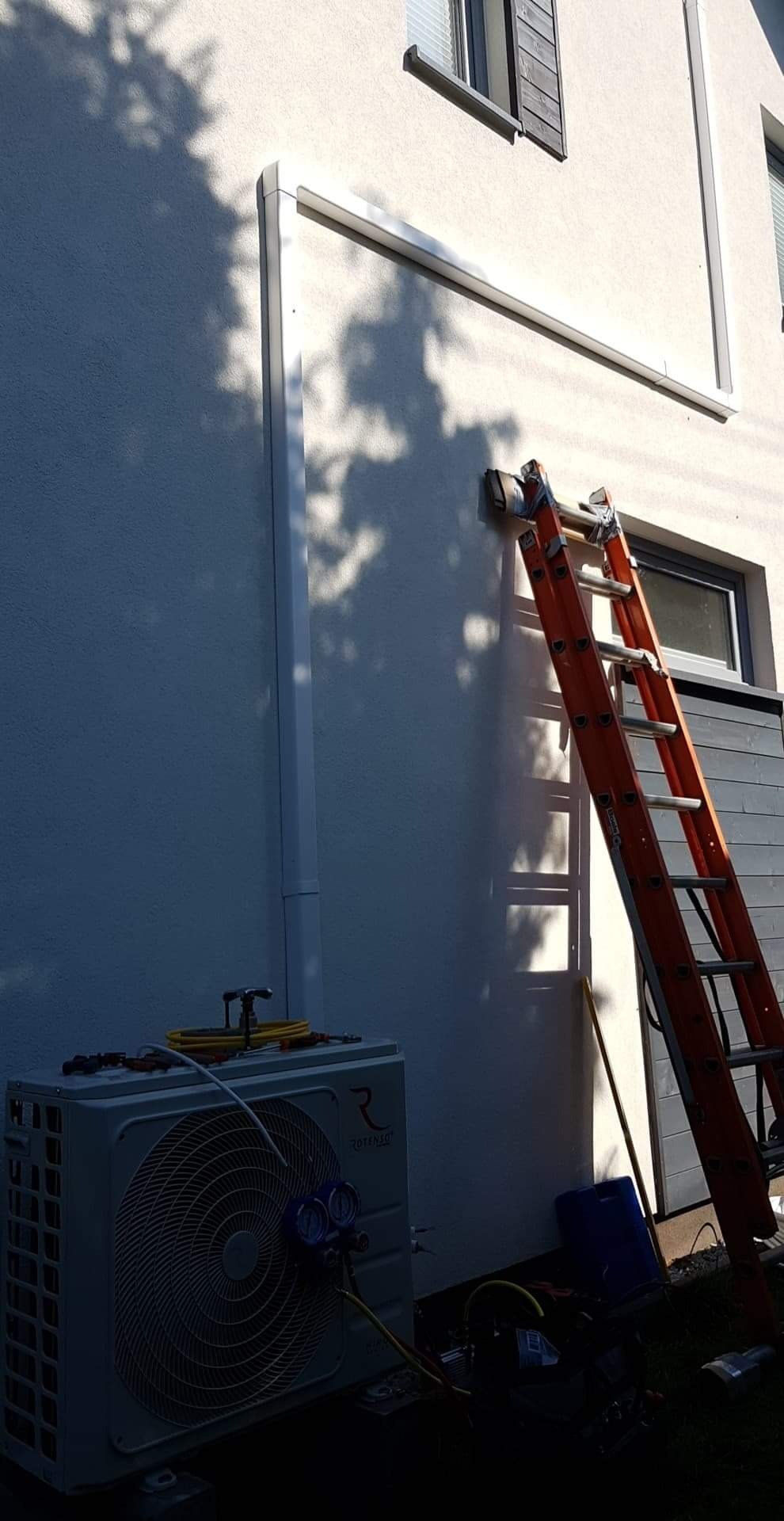 Zewnętrzna jednostka pompy ciepła marki Rotenso, podłączona do manometrów serwisowych, stoi przy ścianie domu z widoczną drabiną opartą o ścianę, cień drzewa na elewacji.