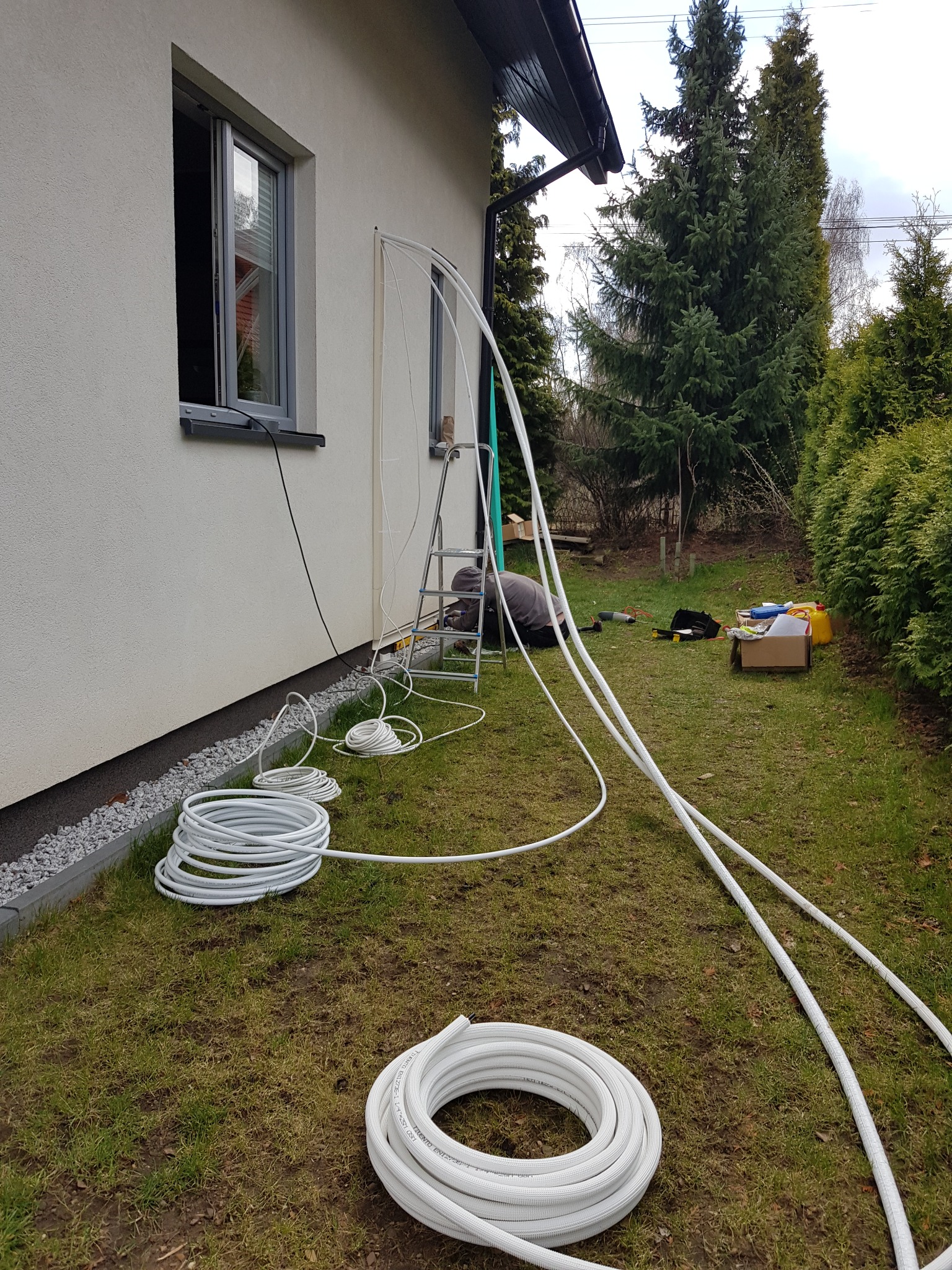 Instalacja białych rur na zewnątrz budynku, widoczny pracownik na drabinie, narzędzia i zwinięte rury na trawie.