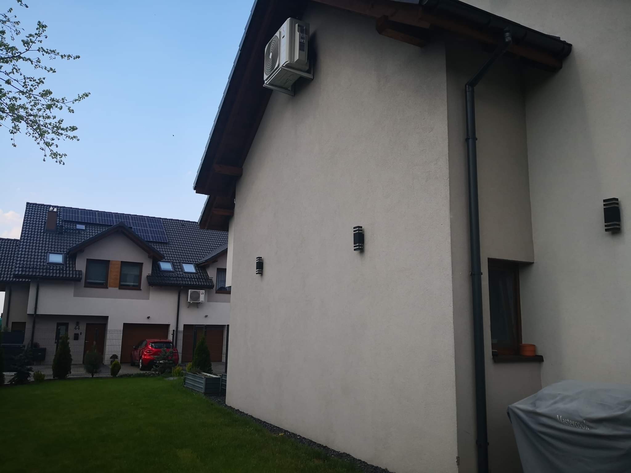 Zewnętrzna jednostka klimatyzacji zamontowana na ścianie domu, widoczna rynna spustowa i oświetlenie elewacyjne, w tle dom z panelami słonecznymi na dachu i czerwony samochód.