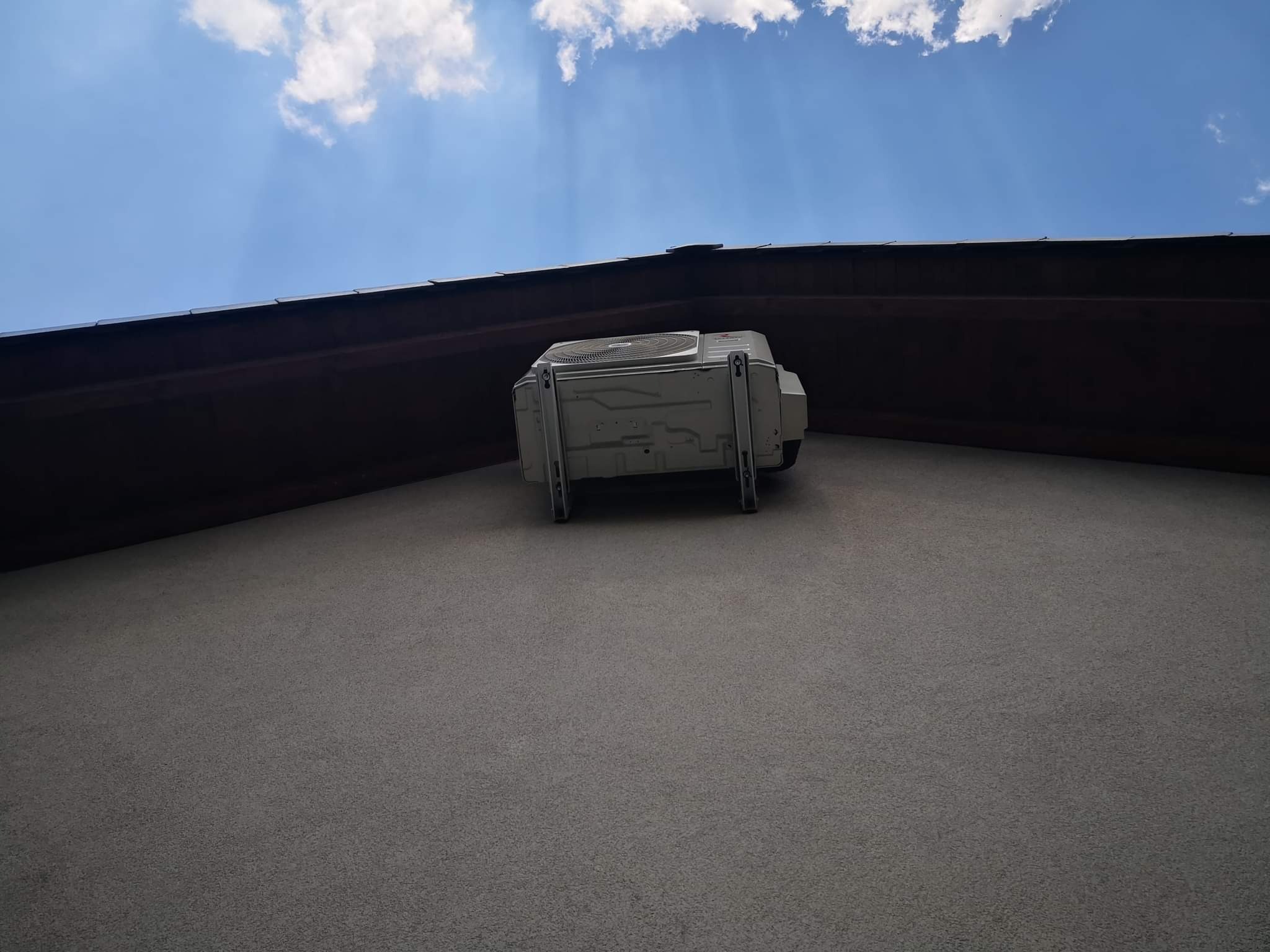 Zewnętrzna jednostka klimatyzacji zamontowana na ścianie budynku pod okapem dachu, widok z dołu w słoneczny dzień.