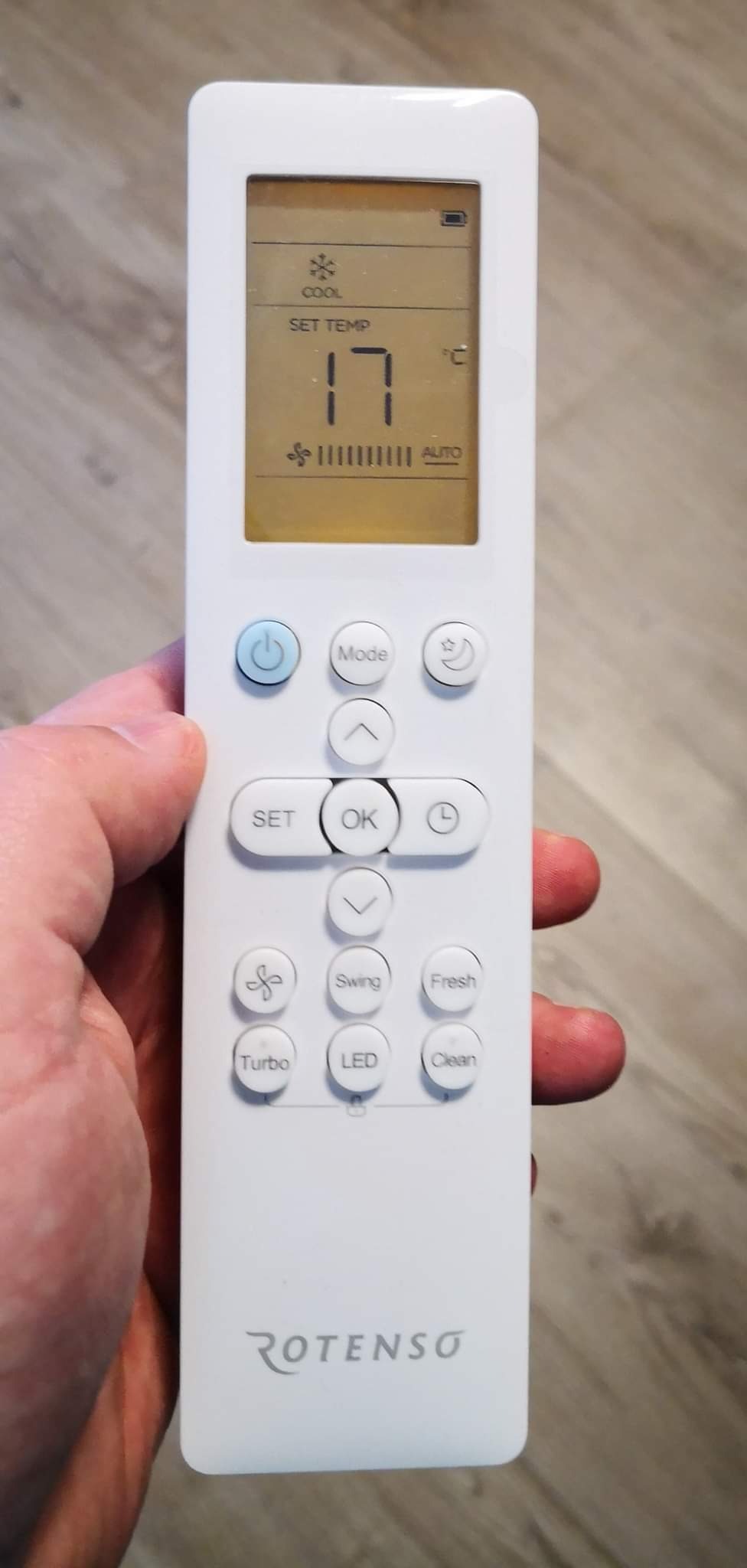 Biały pilot do klimatyzacji marki Rotenso, ustawiony na temperaturę 17 stopni Celsjusza, trzymany w dłoni na tle drewnopodobnej podłogi.