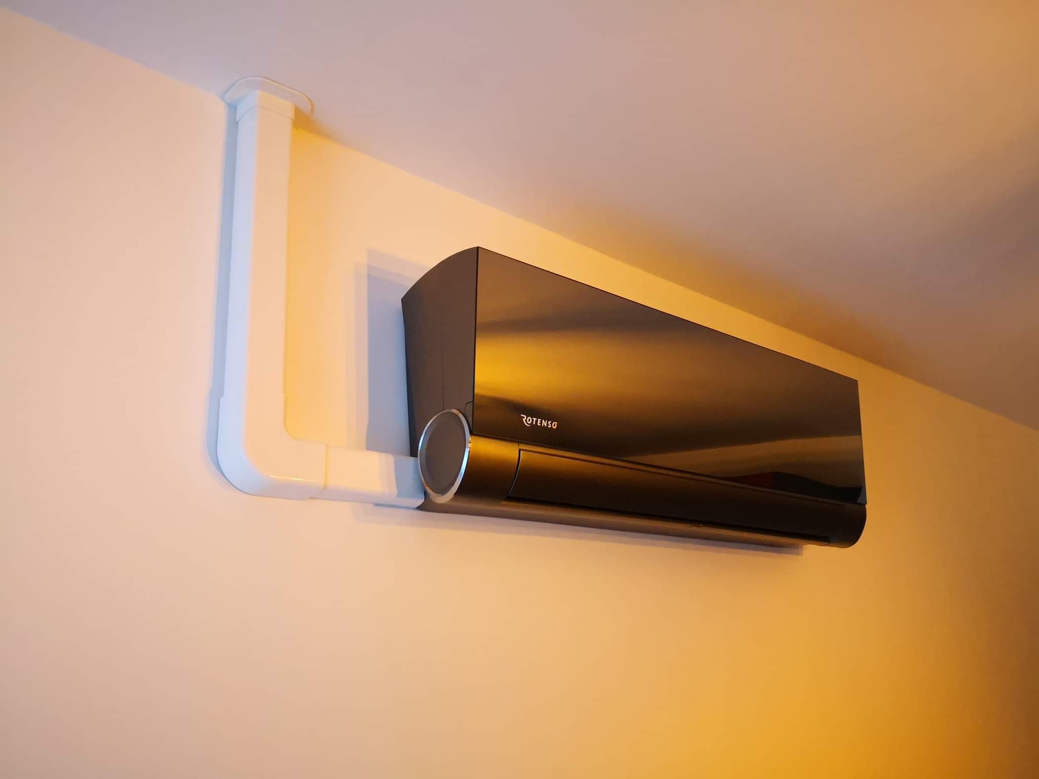 Błyszcząca, ciemna jednostka klimatyzacji marki Rotenso zamontowana na jasnej ścianie z widocznymi białymi rurami instalacyjnymi.