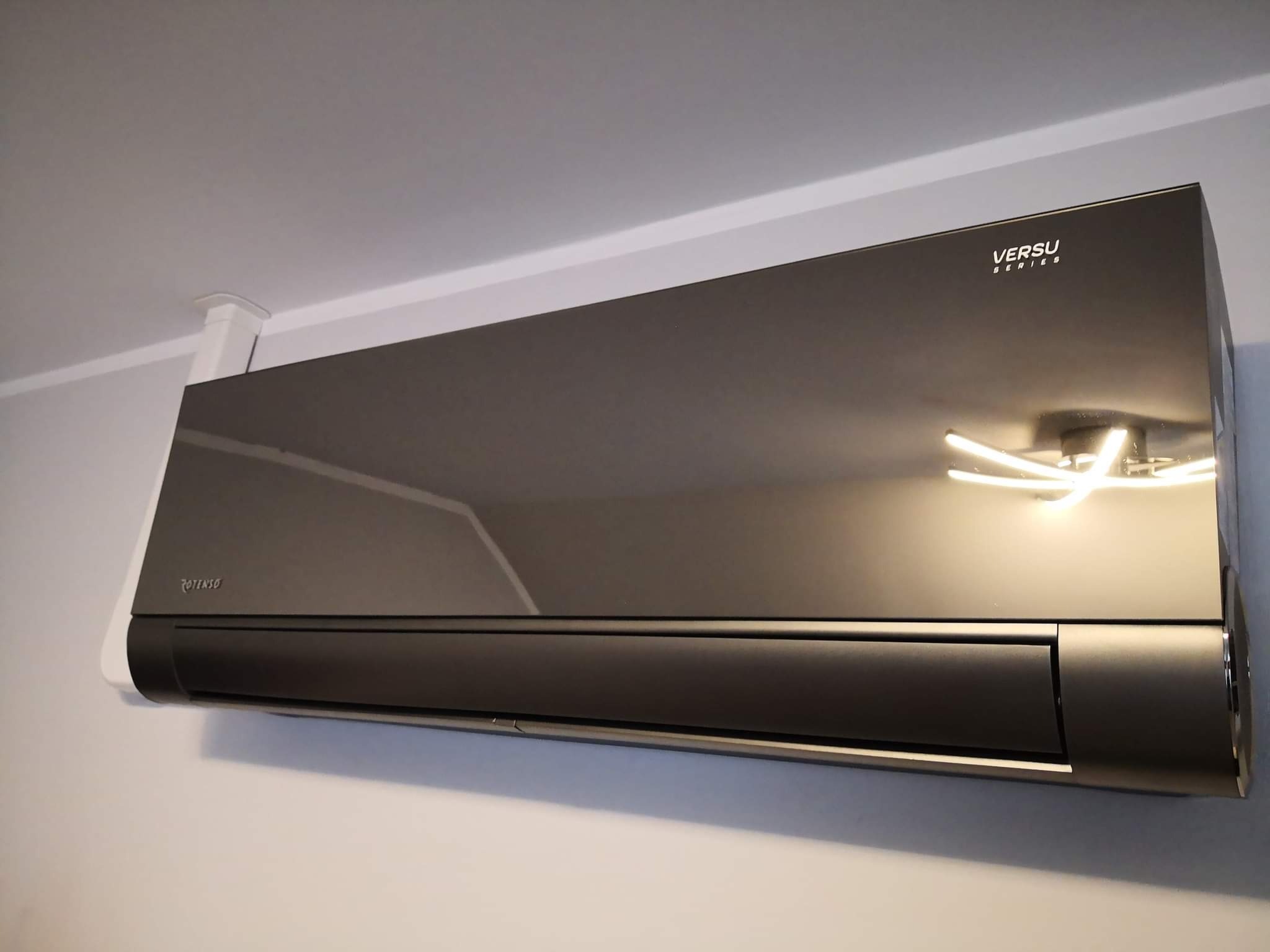 Czarny klimatyzator ścienny marki Rotenso Versu Series zamontowany w biurze, widoczne odblaski oświetlenia na obudowie.