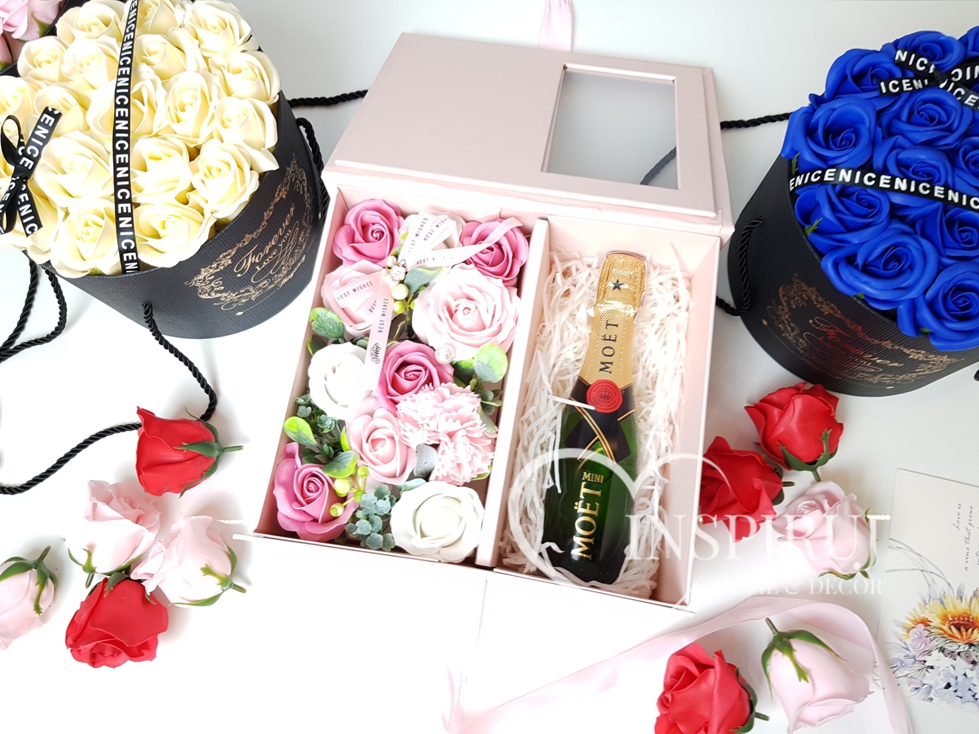 Kompozycja prezentowa: pudełko z różowych i białych mydlanych róż, mała butelka szampana Moët, obok okrągłe pudełka z białych i niebieskich róż, całość na białym tle z rozrzuconymi pojedynczymi...