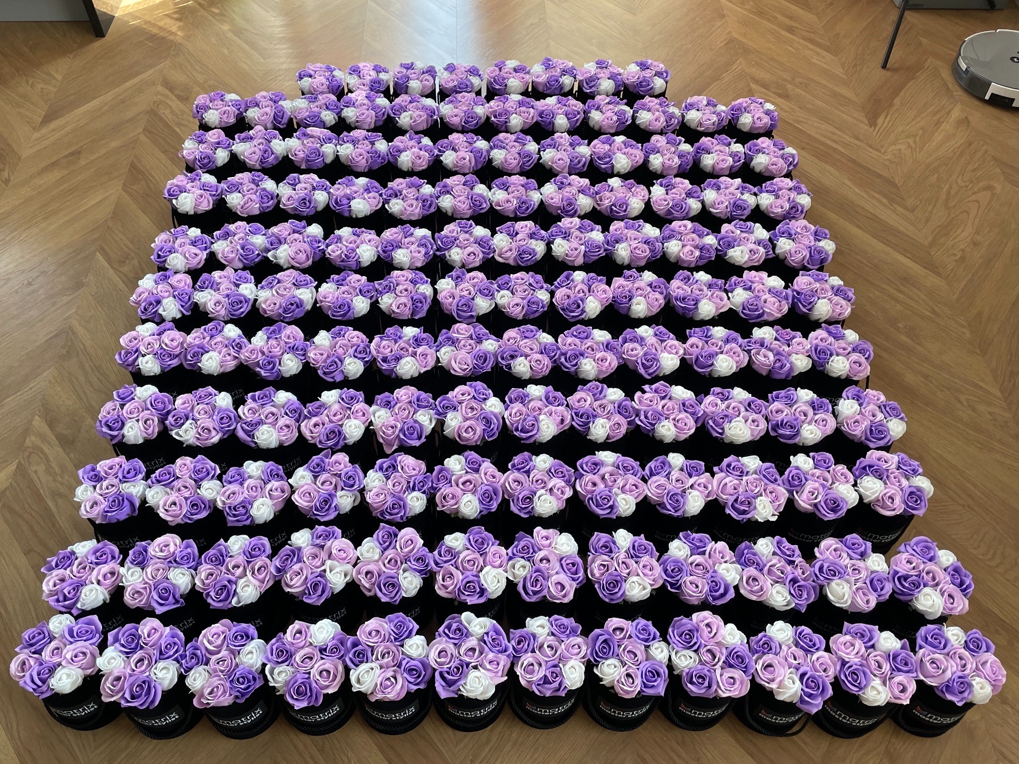 Duża liczba identycznych, małych, okrągłych pudełek z czarnego weluru, wypełnionych kompozycjami z białych i fioletowych piankowych róż, ułożonych w rzędach na podłodze z paneli.