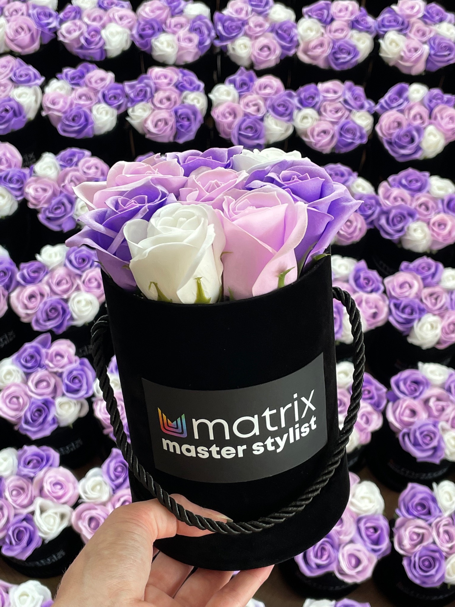 Mały czarny, aksamitny cylinder wypełniony mydlanymi różami w odcieniach bieli, różu i fioletu, trzymany w dłoni na tle wielu identycznych kompozycji; na cylindrze logo Matrix Master Stylist.