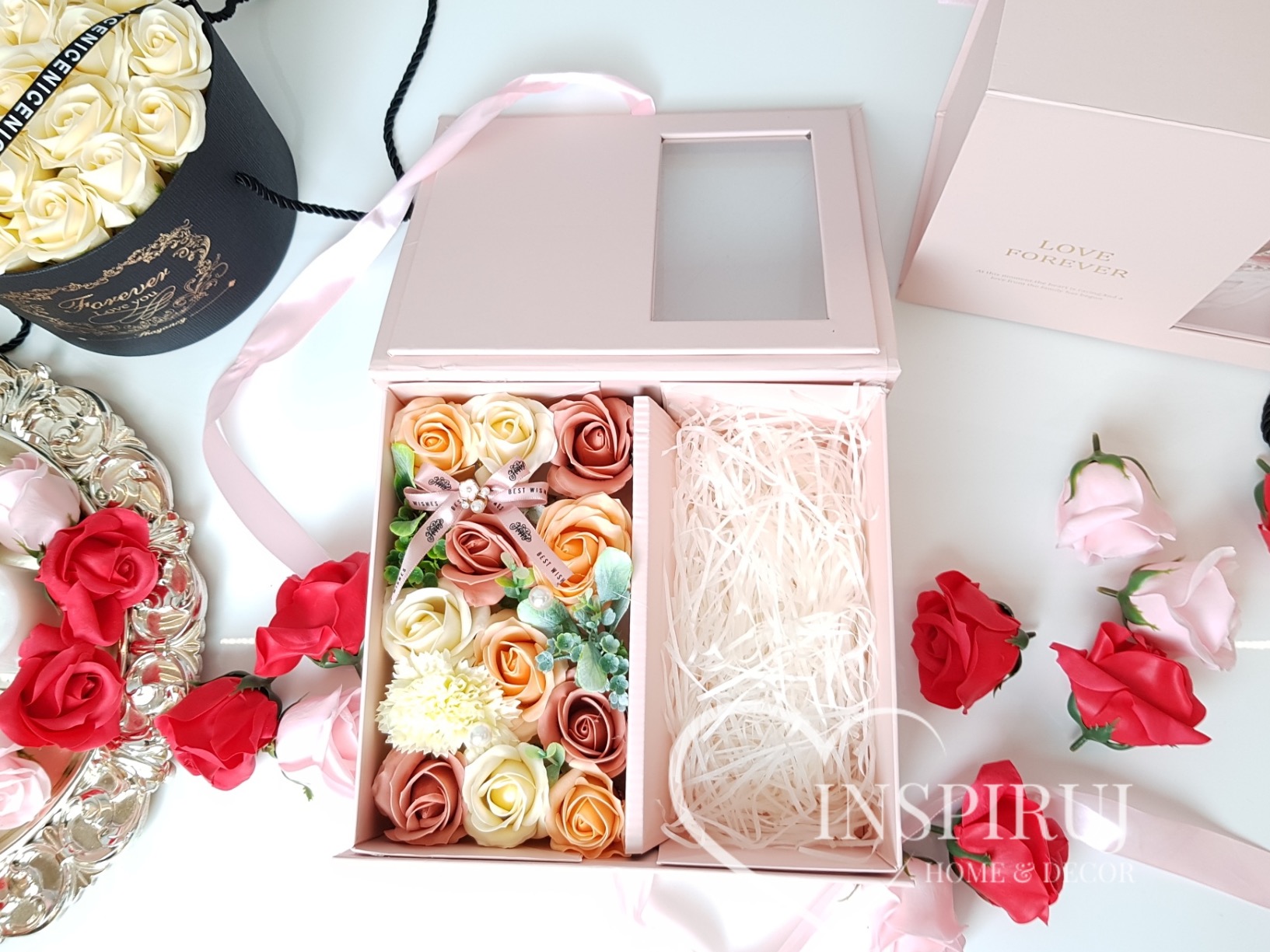 Otwarty, różowy, elegancki box z mydlanymi różami w odcieniach beżu, brzoskwini i bieli, udekorowany wstążką z napisem 'Best Wishes', obok boxu rozsypane pojedyncze róże, w tle okrągły box...