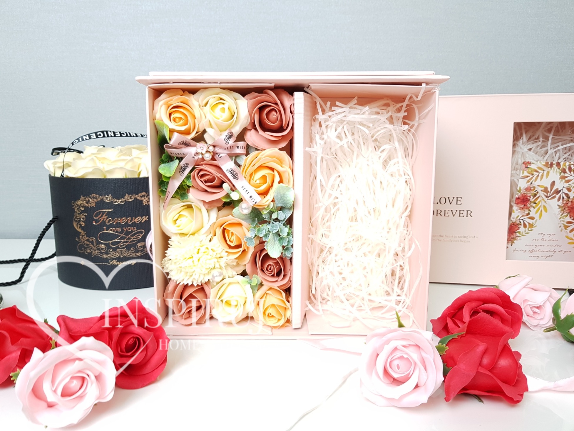Otwarty, jasnoróżowy pudełko prezentowe z przegródkami, wypełnione kwiatami z mydła w odcieniach różu, kremu i brzoskwini, przewiązane wstążką z napisem 'Best Wishes', obok torba prezentowa...