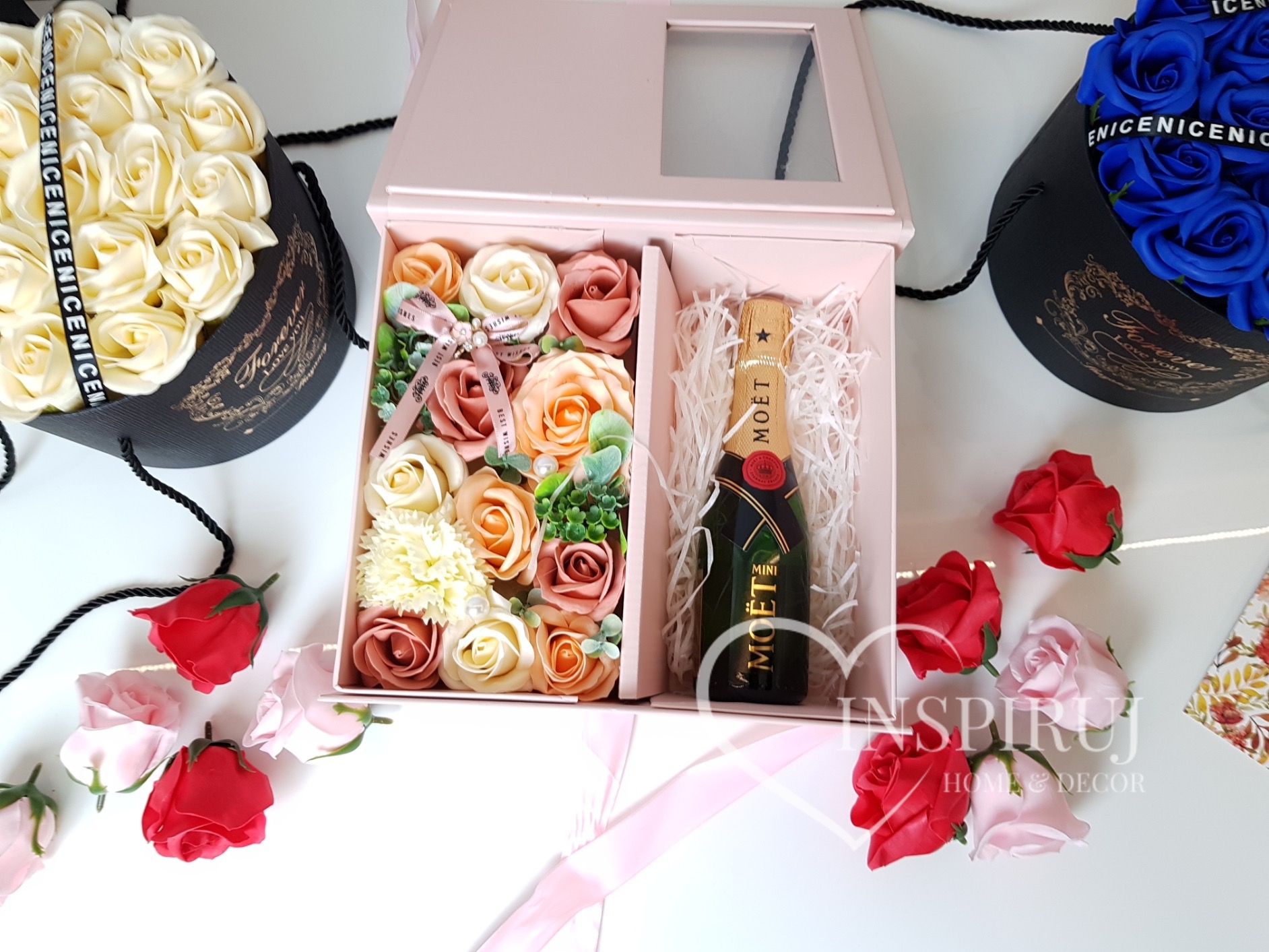 Kompozycja z mydlanych róż w odcieniach beżu, brzoskwini i bieli w jasnoróżowym pudełku z okienkiem, obok mini butelka Moët w białym wypełniaczu, w tle bukiety z białych i niebieskich róż...
